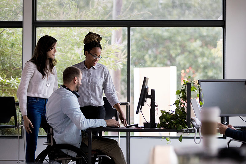 Picture showing 3 people in front of a computer. One man is in a wheel chair.